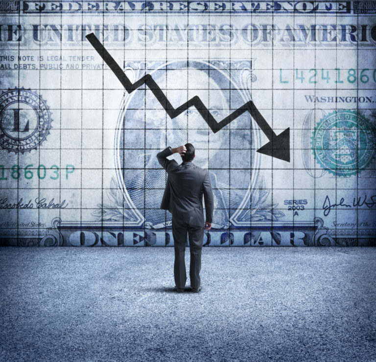 A rear view of a concerned businessman as he places his hand on his head and looks up at a U.S. One Dollar bill coupled with a downward trending arrow and chart on the wall in front of him. This image illustrates the concept of a falling, or weakening U.S. dollar.